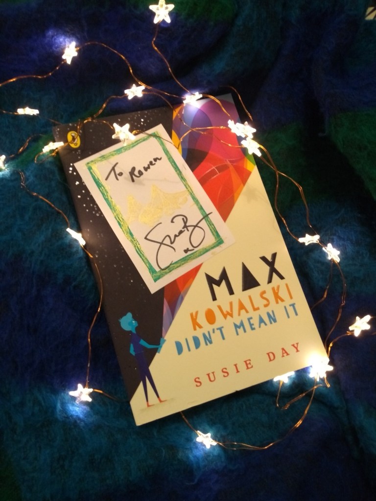 The book Max Kowalski Didn't Mean It by Susie Day, surrounded by star-shaped lights on a dark blue blanket. A bookplate signed by the author is on top.