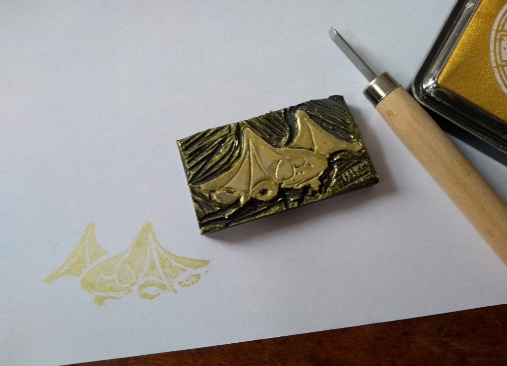 A linocut of a sleeping dragon curled up, with a cutting tool to the right and a print of the linocut in gold ink on white paper