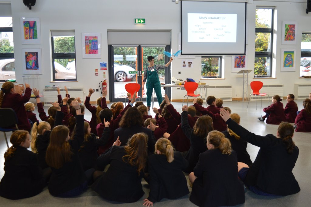 Author Susie Day holds up a pair of fluffy blue ears in front of an audience of 10+ children, most of them holding up their hands to answer a question.